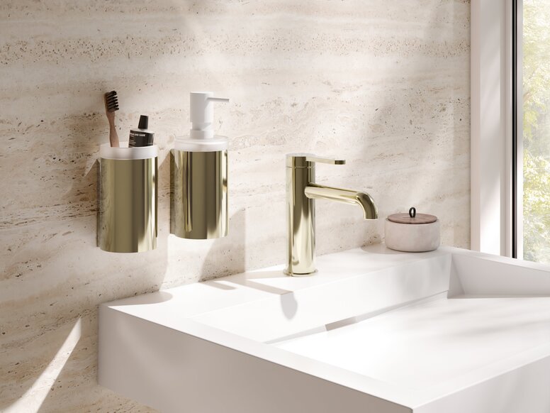 A brass-coloured PVD toothbrush holder and soap dispenser next to a tap on a white washbasin against a beige wall