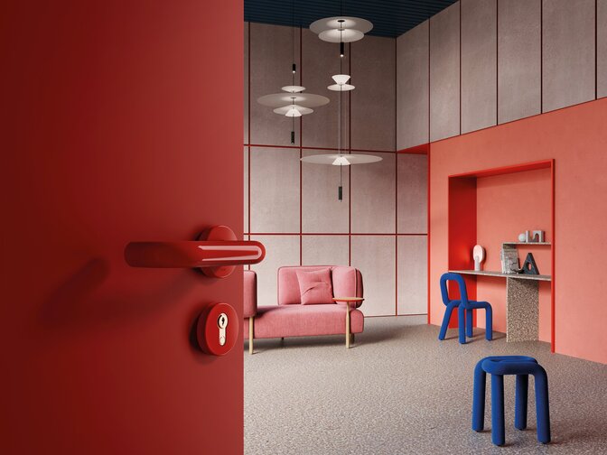 A red door handle with a rose and keyhole on a red door; behind it, a living room with a workspace