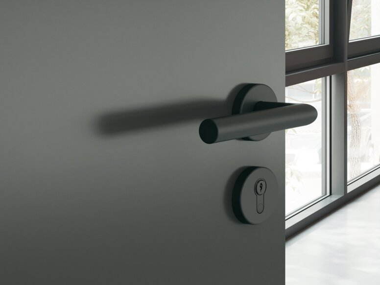 A dark grey powder-coated door handle with a round rose on a dark grey door; behind it is a study with a desk and chair