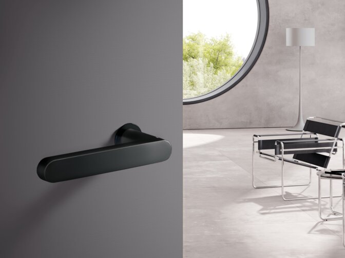 A black powder-coated door handle fitted to a grey door; behind it, a bright room with two chairs