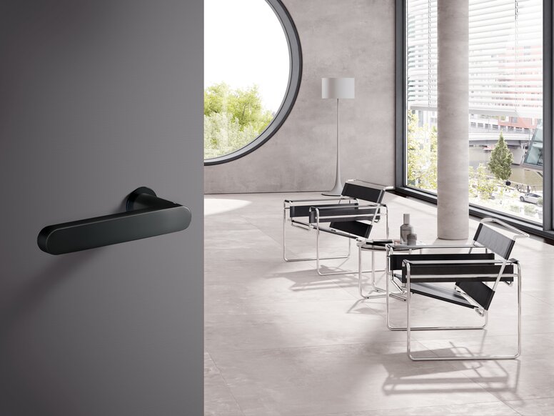 HEWI Range 270 lever handles A black powder-coated door handle fitted to a grey door; behind it, a bright room with two chairs