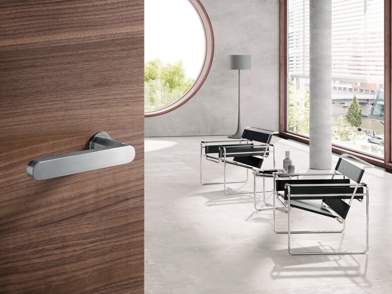 A brushed stainless steel door handle on a wooden door; behind it, a room with black chairs on a grey floor and a row of windows