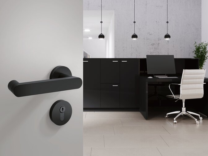 A black door handle with a round rosette plate fitted to a light grey door; behind it is a reception area with a counter and a chair