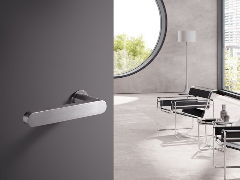 HEWI Range 270 lever handles A stainless steel door handle on a grey door; behind it, a bright room with chairs