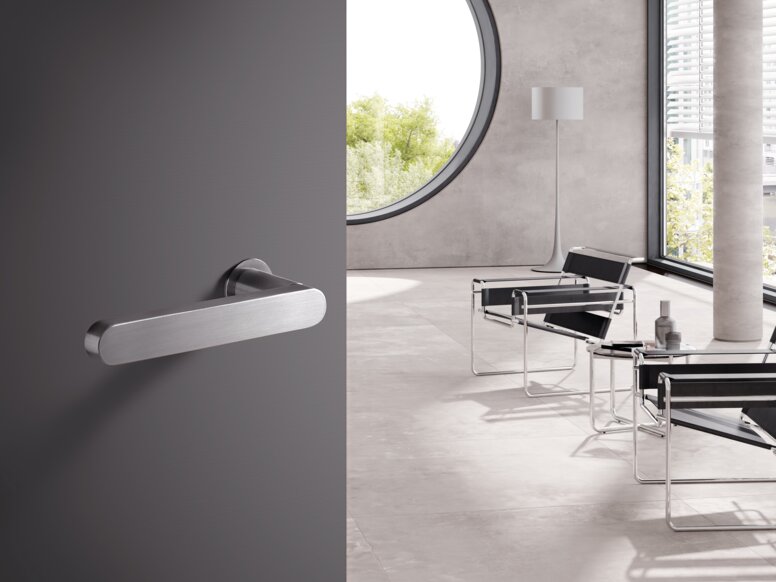 A stainless steel door handle on a grey door; behind it, a bright room with chairs