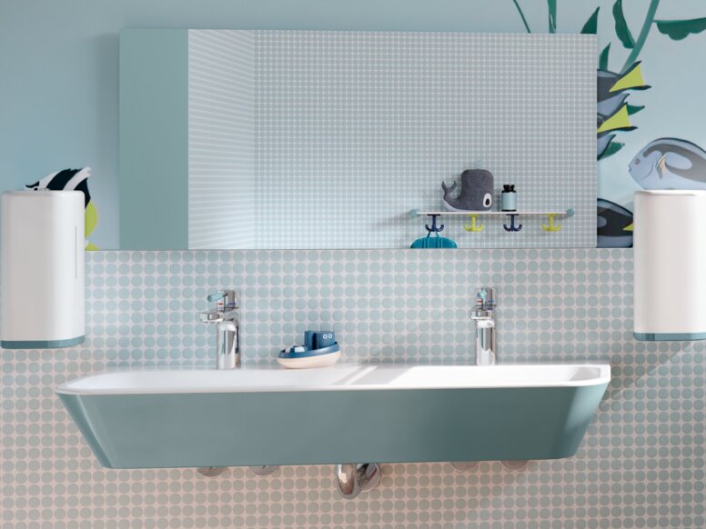 A blue children’s washbasin made of mineral composite, with soap dispensers and a mirror above it, next to which is a WC area