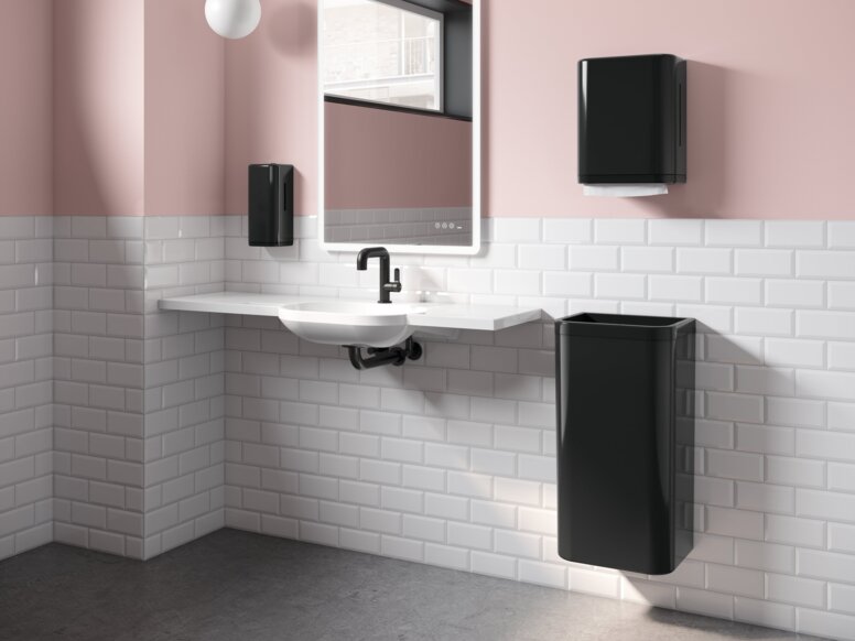 A white washbasin with a black paper towel dispenser and wastebin next to it, in a bathroom with white tiles and pink walls