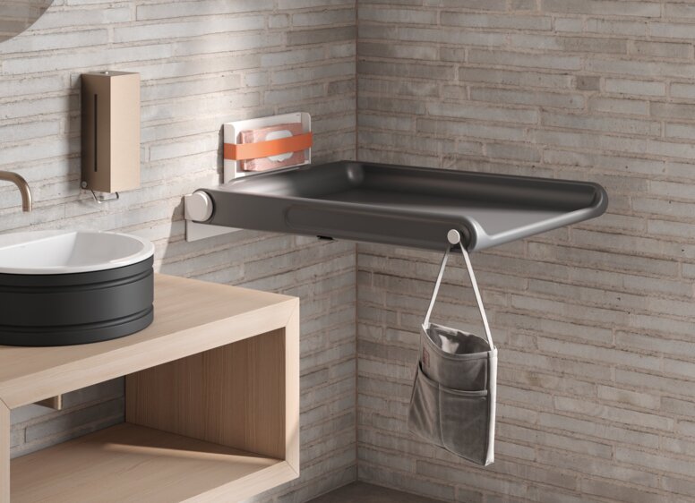 HEWI Range 480 Baby Changing Table A hinged baby changing table with a side pocket for storage and a hook, mounted on a light-coloured brick wall