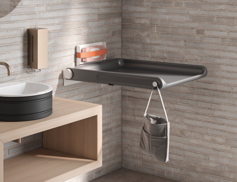 A hinged baby changing table with a side pocket for storage and a hook, mounted on a light-coloured brick wall