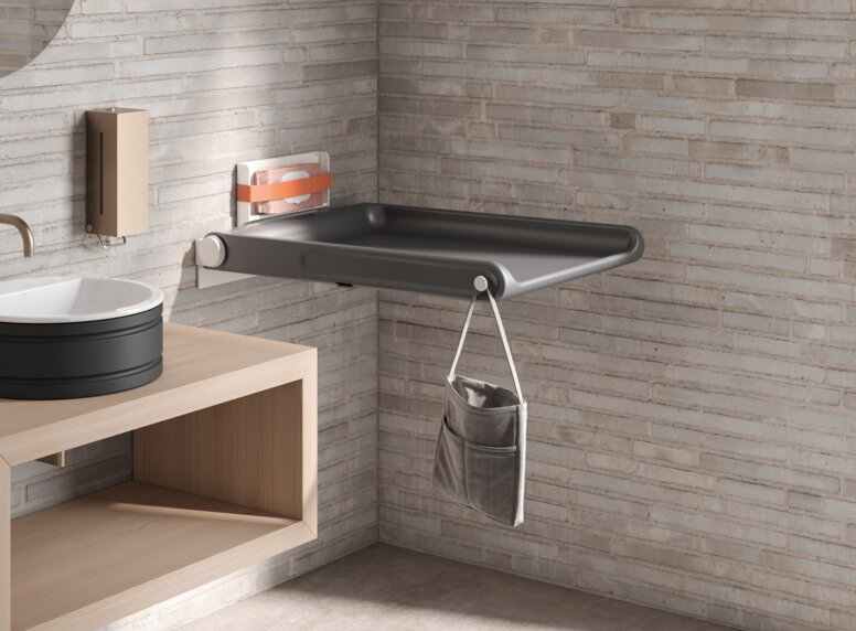 HEWI Serie 480 Babywickeltisch Klappbarer Babywickeltisch mit Seitentasche zur Aufbewahrung und einem Haken an heller Ziegelwand montiert
