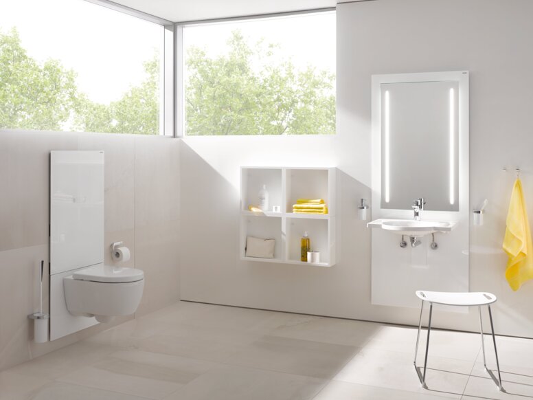 A white WC module in a bright bathroom with an illuminated mirror above the washbasin, in front of which stands a stool with a white seat