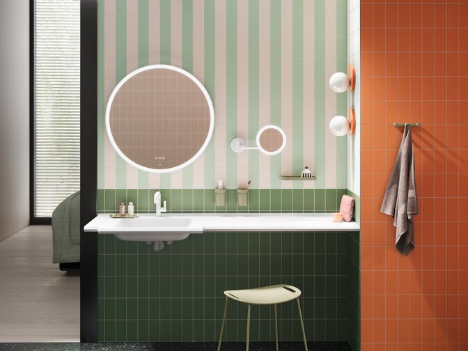 Washbasin with green fittings, a round mirror, cosmetic mirror and stool against a green-and-white striped wall and orange tiles
