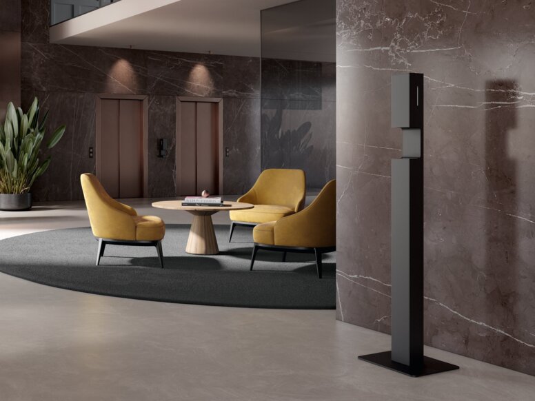A free-standing black powder-coated hand sanitiser stand in a reception area in front of a brown marble wall