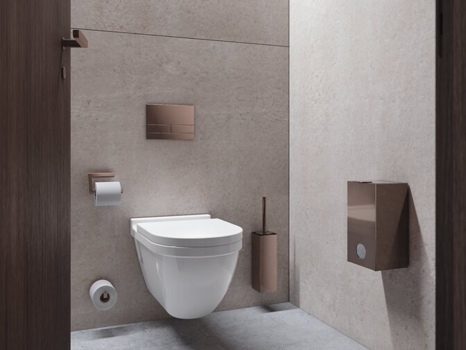 Wall-hung WC with toilet brush and toilet roll holder, featuring a push-button flush in a copper finish, mounted on a beige concrete wall