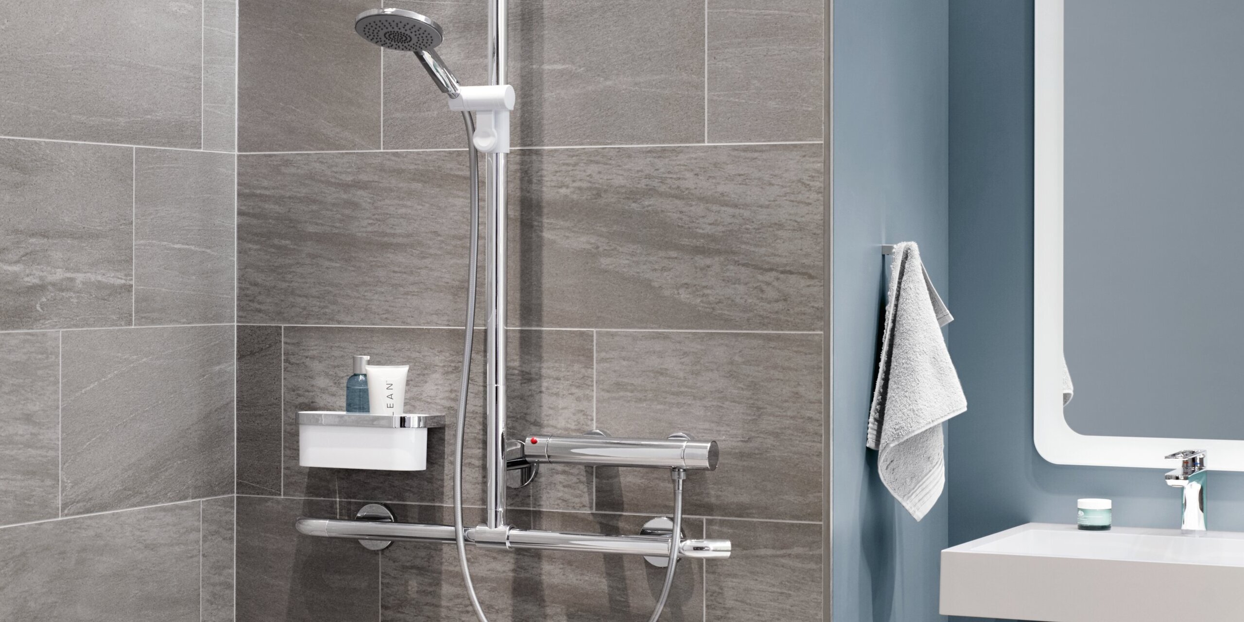 A hinged shower seat with a support rail, a thermostatic mixer and a shower head against a grey tiled wall, next to which is a washbasin area against a blue wall