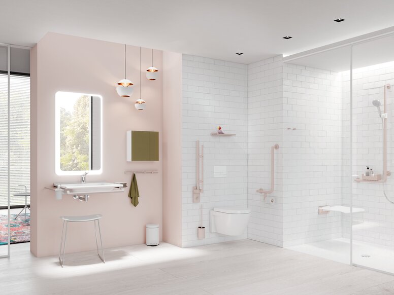 Bright bathroom with a pale pink feature wall, illuminated mirror above a white washbasin, wall-hung toilet and level-access shower with white wall tiles and light-coloured grab rails